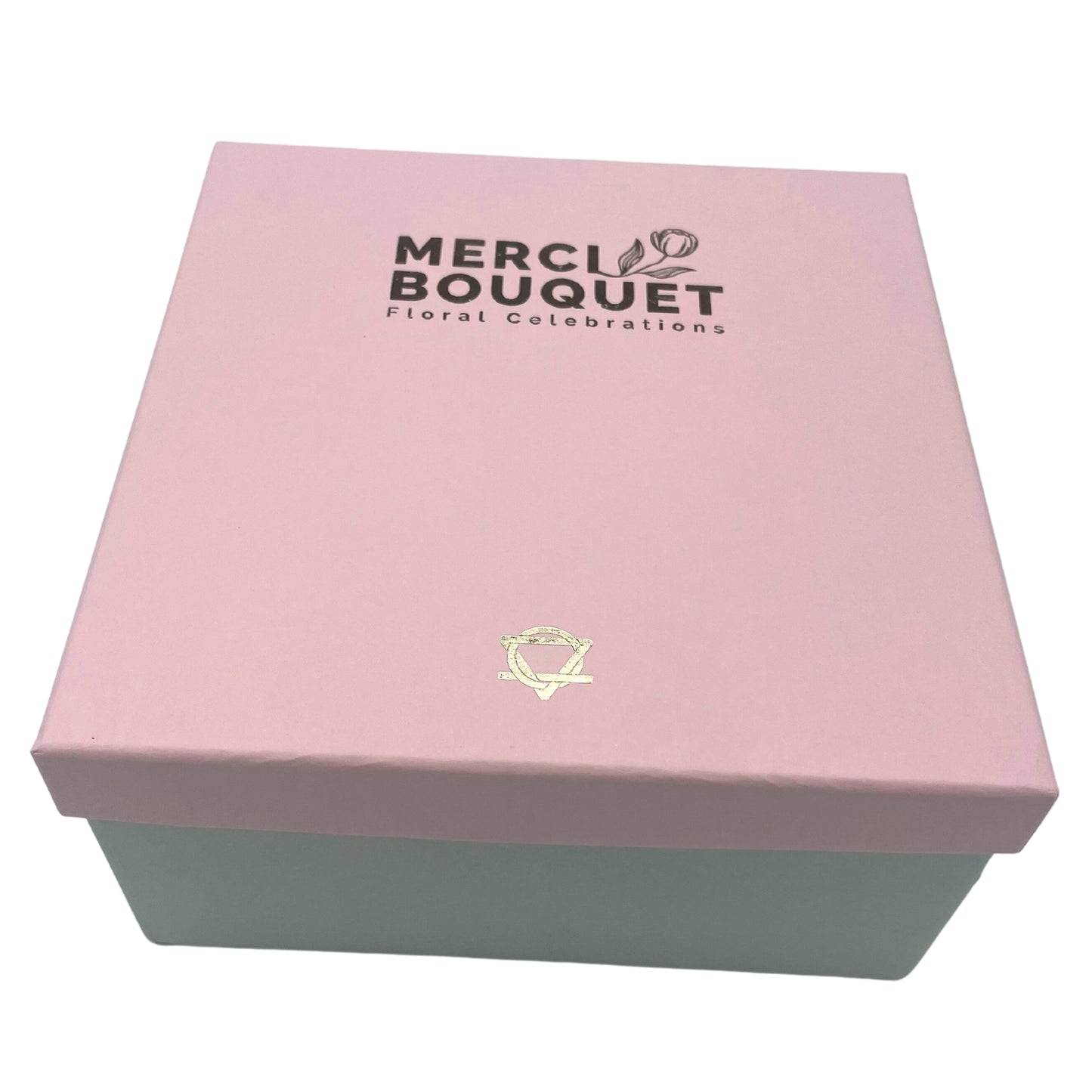 A pink box with a light green base sits on a surface. The lid displays "MERCI BOUQUET Floral Celebrations". A gold symbol is centered below the text.