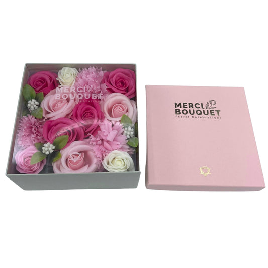 A box of pink and white soap flowers sits atop a light grey box. The flowers are arranged in a square shape. The larger box is pink and says "MERCI BOUQUET Floral Celebrations" on it.