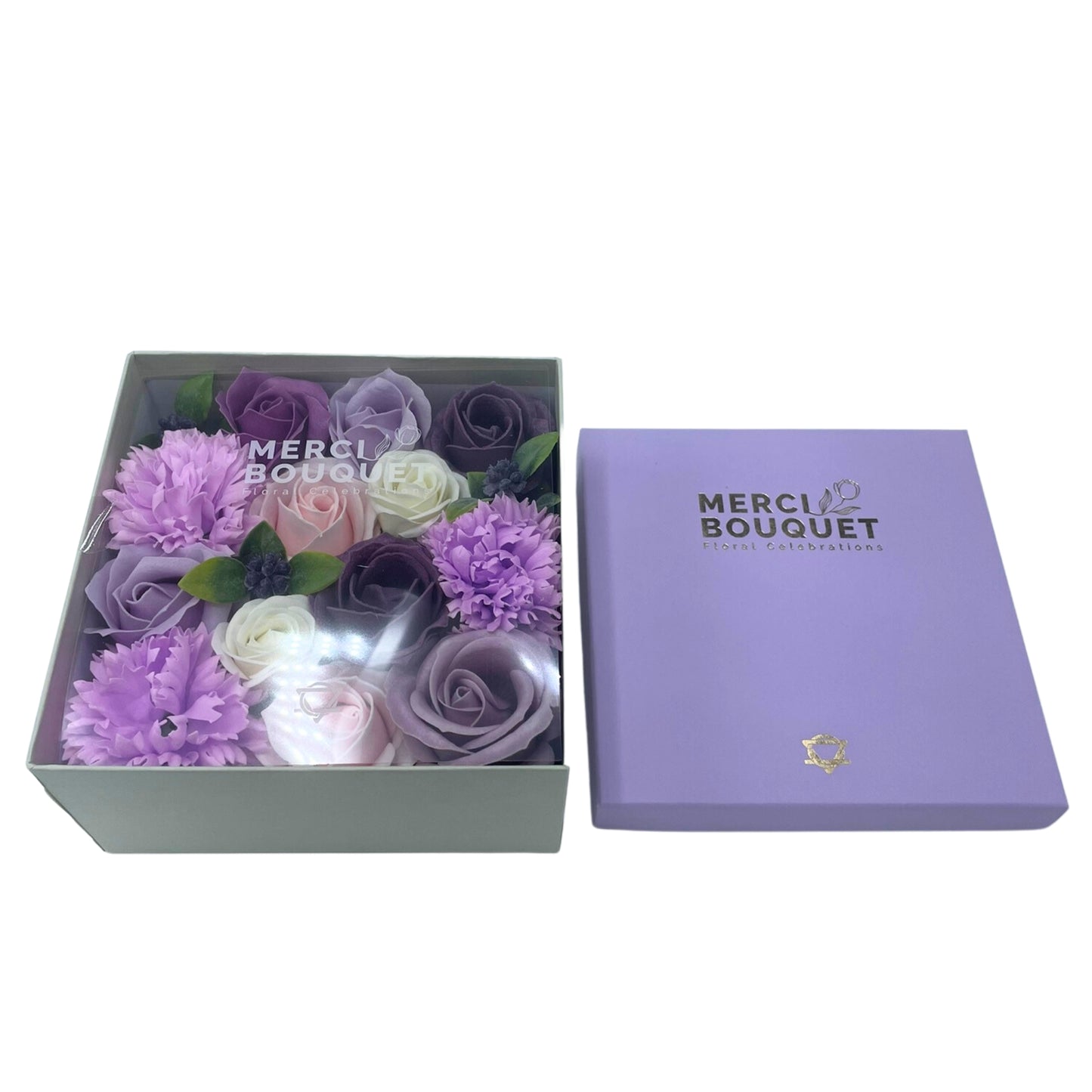 A bouquet of purple and pink soap roses and carnations sits in a light grey box. The box is next to a lavender-colored box with "MERCI BOUQUET Floral Celebrations" printed on it.