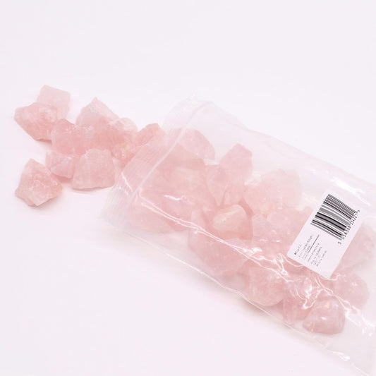 Pink rose quartz chunks spill from a clear plastic bag onto a white surface. The bag has a label with product information and a barcode.