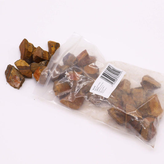 Brown tiger eye stones are spilling from a clear plastic bag onto a white surface. The bag has a label with barcode and text: "SKU:YA Raw Crystal Chips. Size: Small. Weight: 7 oz (198g). UPC: 706838332274. Type: Tiger Eye."