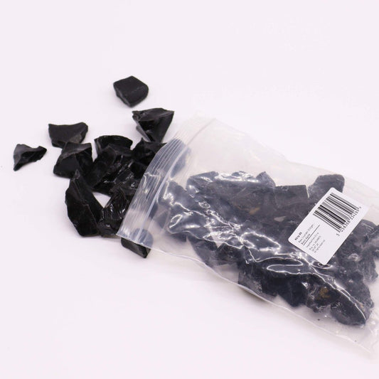 Black obsidian pieces spill from a clear plastic bag onto a white surface. The bag's label reads: KC725, Size: 0.50x0.75in, Black Obsidian, Imported from China, 5117-C 0516881, 8117-K 0516881, & Organics.