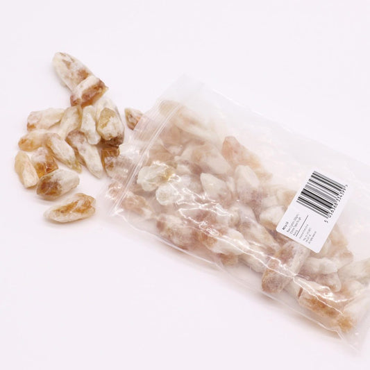 Many small, light brownish-orange, pointed crystals spill from a clear plastic bag onto a white surface. The bag's label reads: "RCry-10 Points Raw Crystals (500gm) Corine Topaz Rough Imported from China".