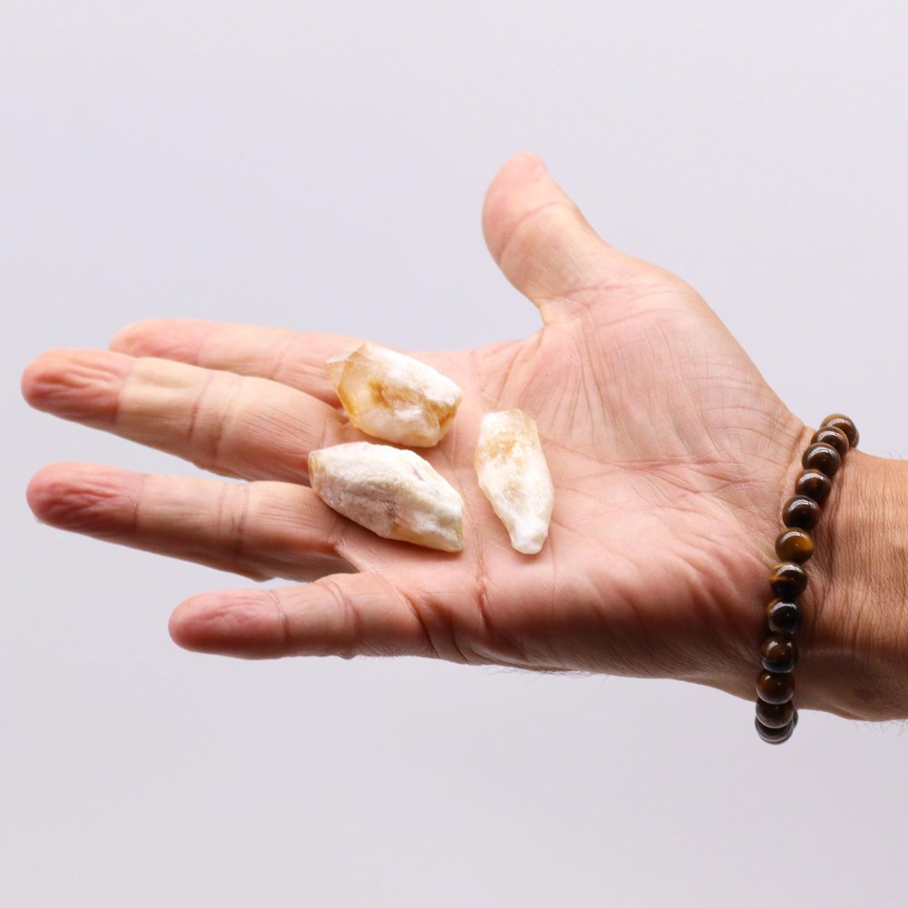Three small, pale yellow-white stones rest on a person's open palm, which is adorned with a brown beaded bracelet. The hand is shown against a plain white background.