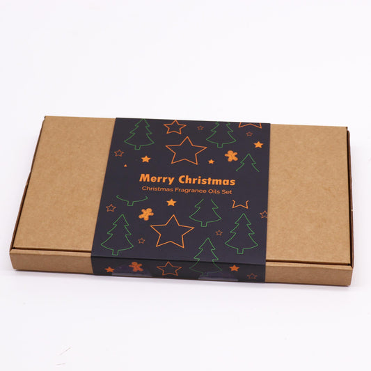 A cardboard box sits on a white surface. It displays a festive label reading "Merry Christmas Christmas Fragrance Oils Set," featuring green trees, orange stars and gingerbread men.