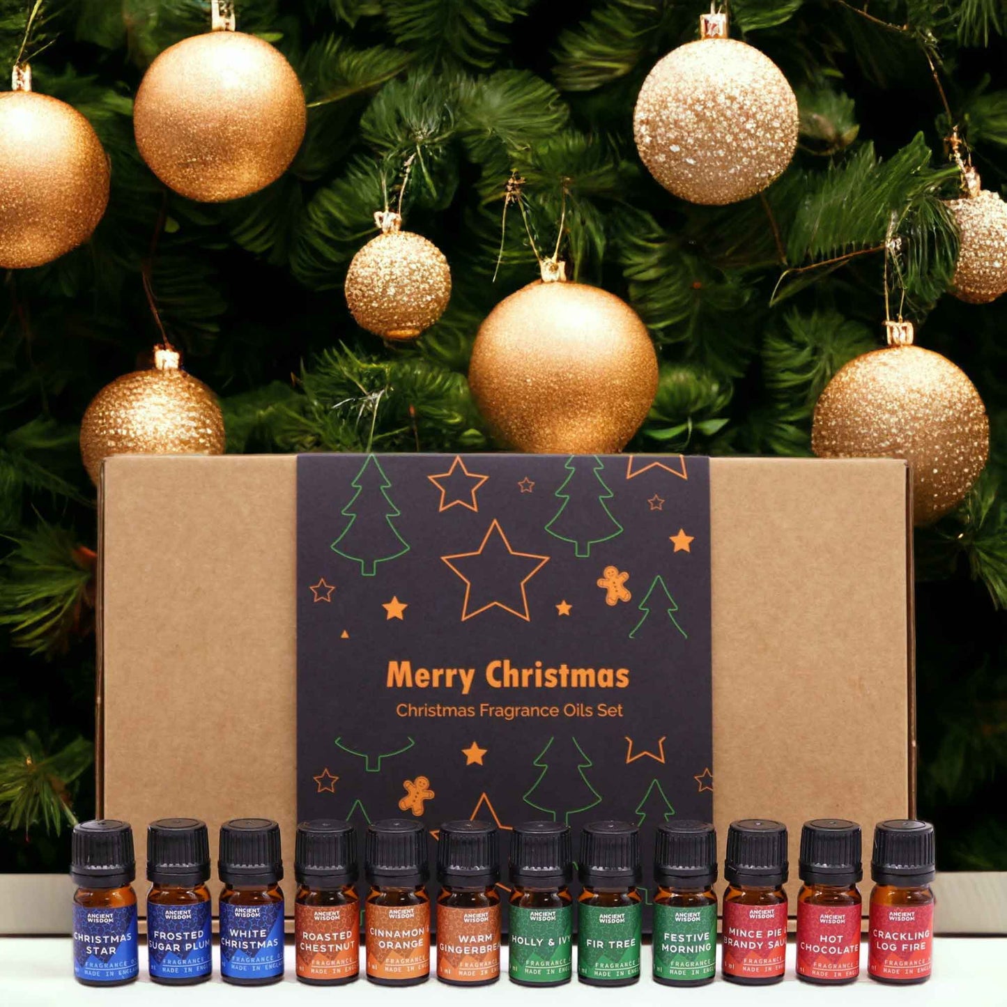A box of twelve small bottles of Christmas-themed fragrance oils sits in front of a decorated Christmas tree. Merry Christmas Christmas Fragrance Oils Set.