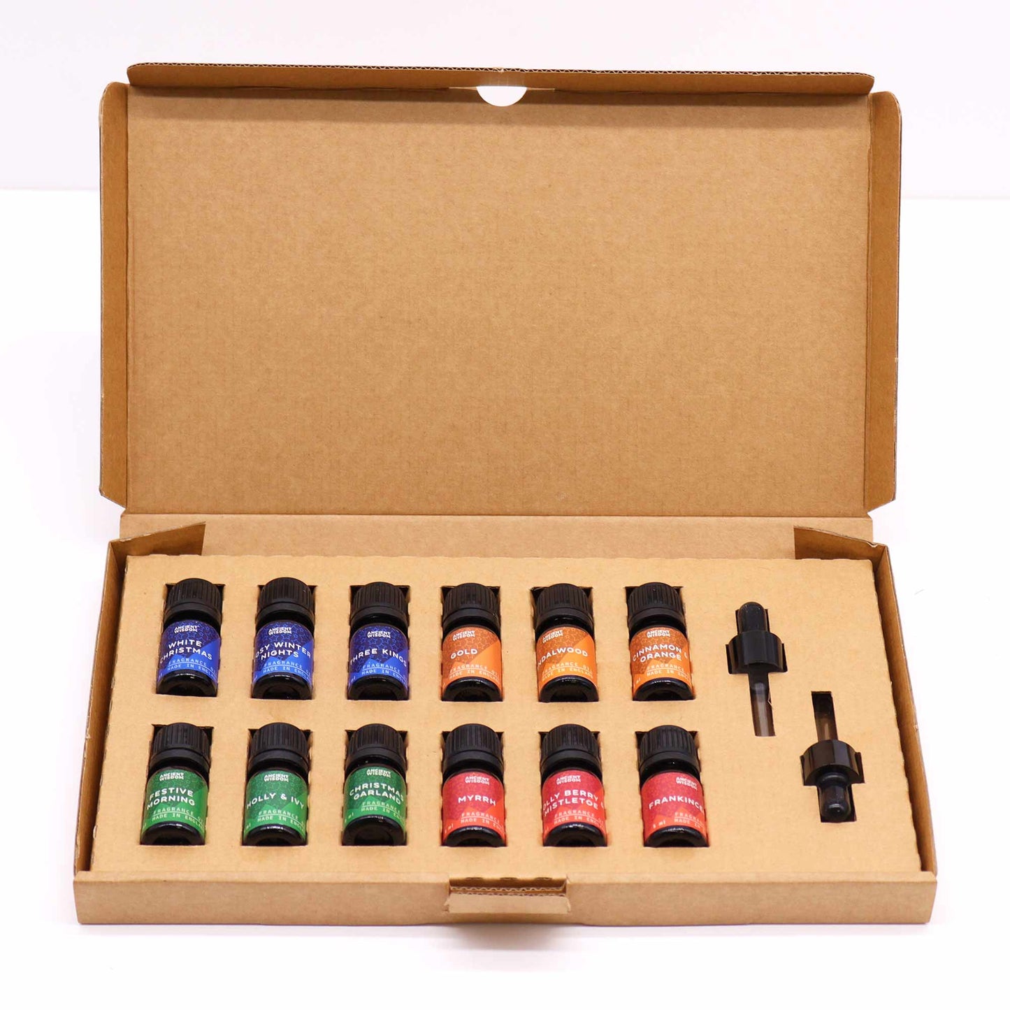 Twelve small bottles of essential oils rest inside a cardboard box; a dropper is included. Each bottle is labeled with a Christmas-themed scent name and “Ancient Wisdom Fragrance, Made in England”.