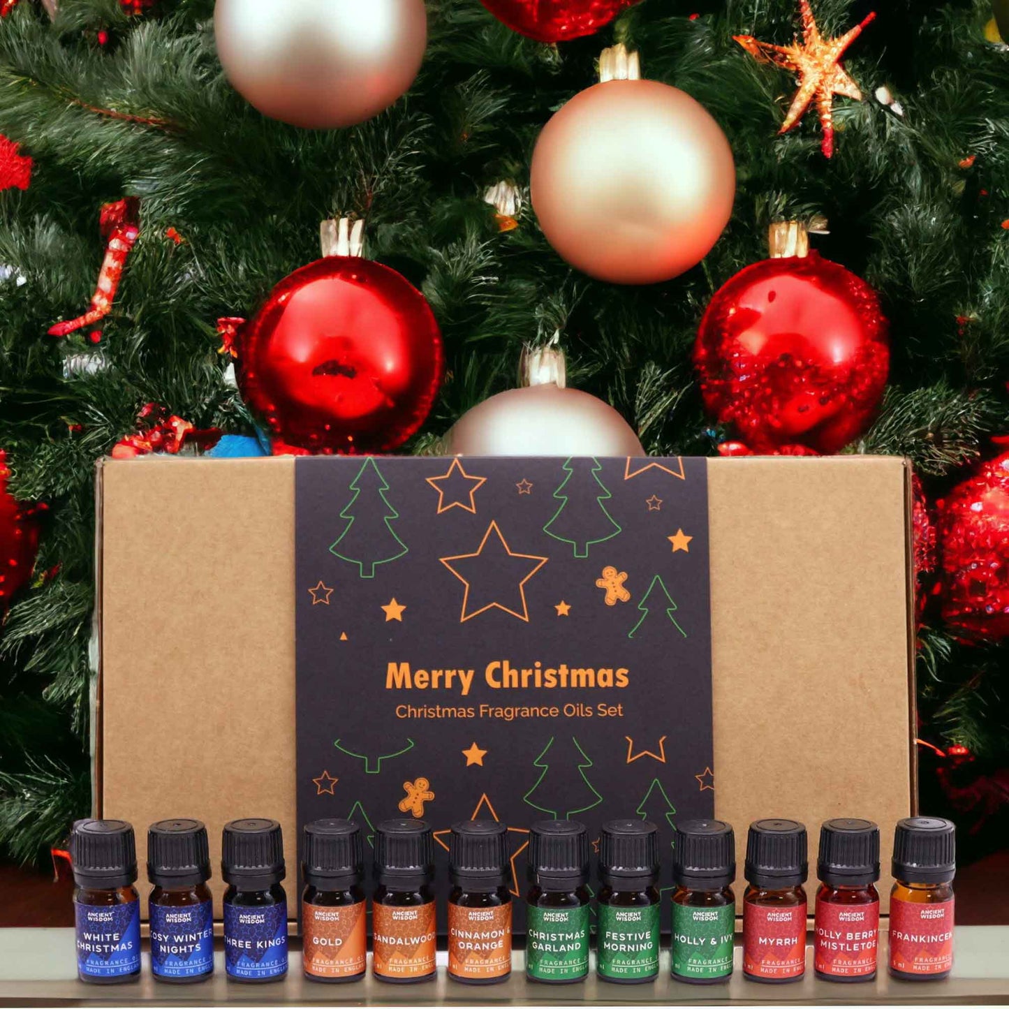 A Christmas fragrance oil set rests in front of a decorated Christmas tree. The box displays "Merry Christmas, Christmas Fragrance Oils Set". Ten small bottles of various scents are included.