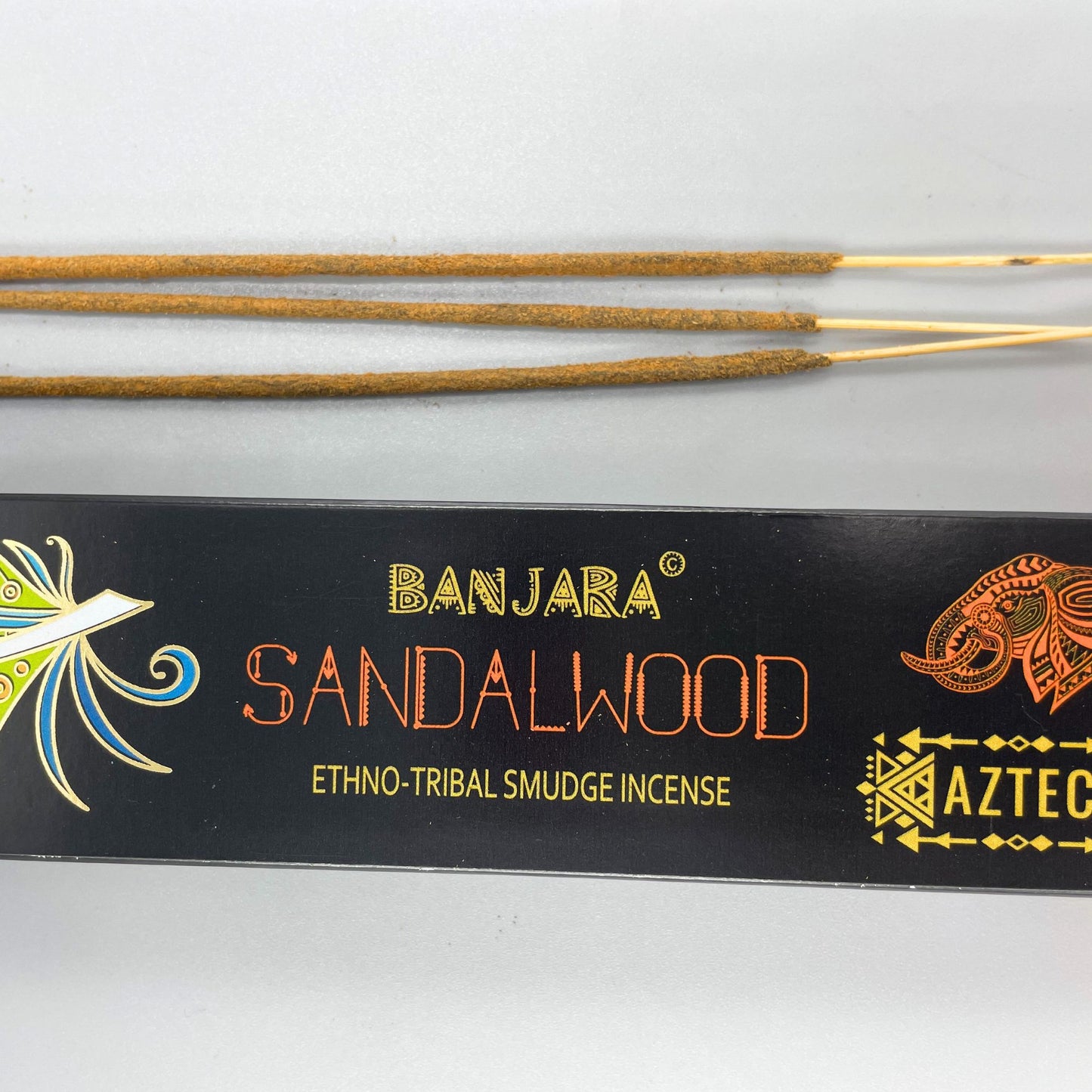 Incense sticks lying on a surface, with a packaging box below. Text reads "BANJARA SANDALWOOD ETHNO-TRIBAL SMUDGE INCENSE" and "AZTEC." The box features decorative tribal patterns.