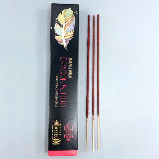 Incense sticks rest next to a black box labeled "BANJARA DRAGON BLOOD ETHNO TRIBAL SMUDGE INCENSE," adorned with a colorful feather design and Aztec motifs, on a light surface.