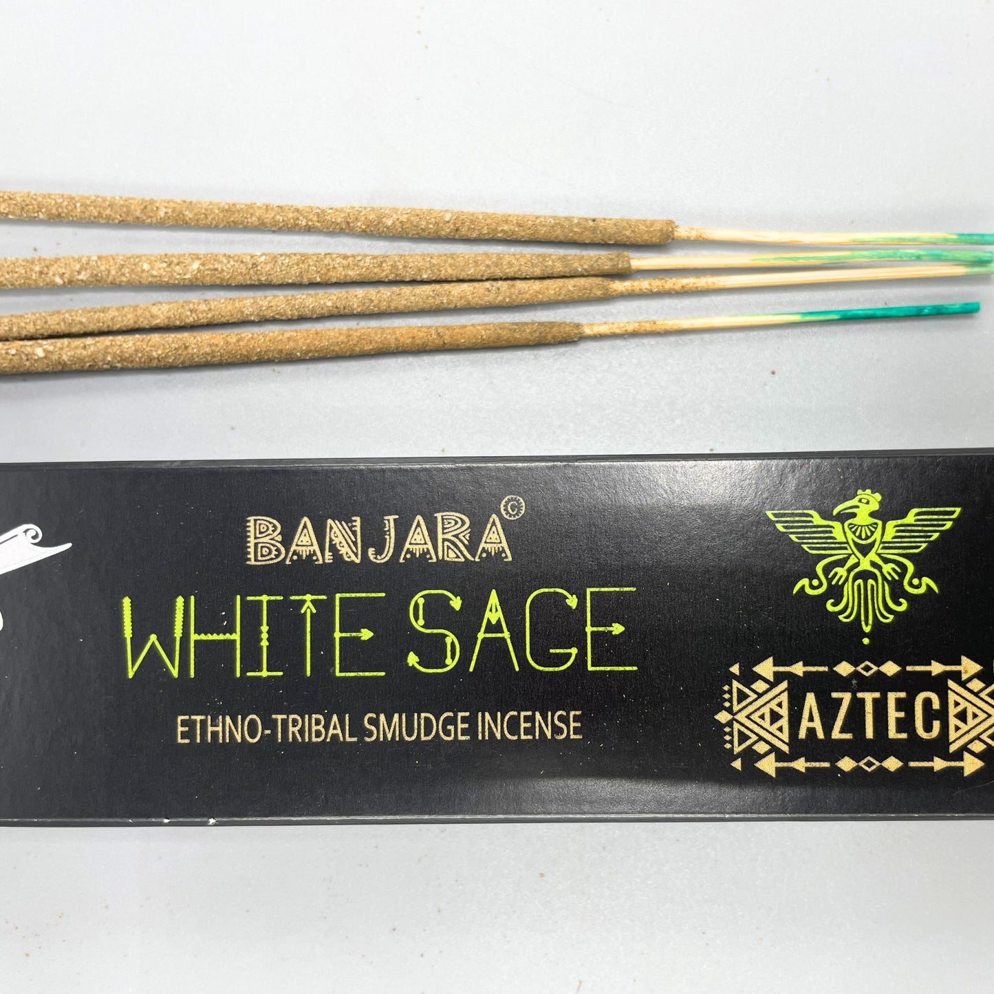 Incense sticks rest next to black packaging labeled "BANJARA WHITE SAGE ETHNO-TRIBAL SMUDGE INCENSE AZTEC," featuring tribal designs and a stylized bird emblem, set against a plain background.
