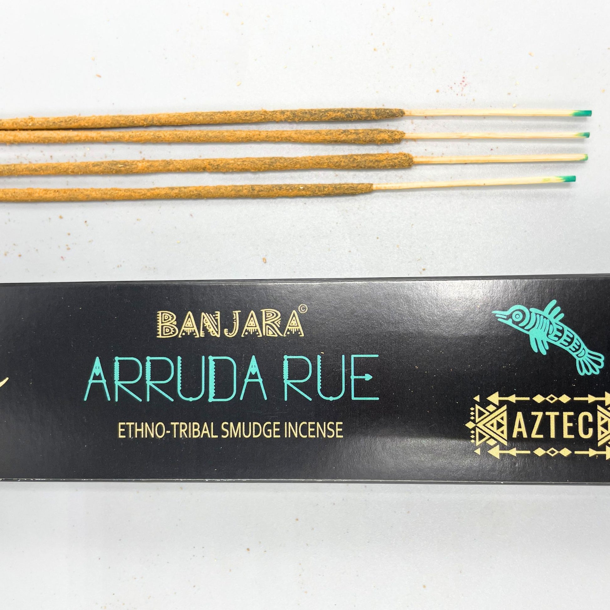 Incense sticks lay beside a black box labeled "BANJARA ARRUDA RUE ETHNO-TRIBAL SMUDGE INCENSE AZTEC," featuring tribal graphics and a turquoise fish design on a light surface.