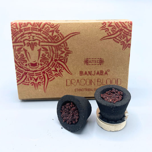 Two black incense cones, with reddish interiors, rest near a brown box featuring a red tribal bull design. Text reads: "AZTEC BANJARA DRAGON BLOOD, ETHNO TRIBAL SMOKE."