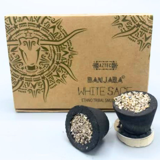 A brown box labeled "Banjara White Sage Ethno Tribal Smudges" sits behind two black incense cones filled with white sage pieces, resting on a light surface.