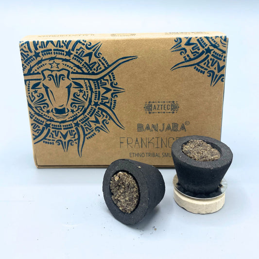 Two dark, conical smudge cups filled with a granular substance sit in front of a cardboard box. The box features tribal-style bull imagery and text: "AZTEC, BANJARA, FRANKIN, ETHNO TRIBAL SMUDGE."