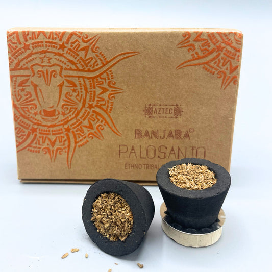Two dark incense cones filled with crushed palo santo rest on a light surface. Behind them, a brown box features an Aztec-style design and text: "BANJARA® PALOSANTO ETHNO TRIBAL."