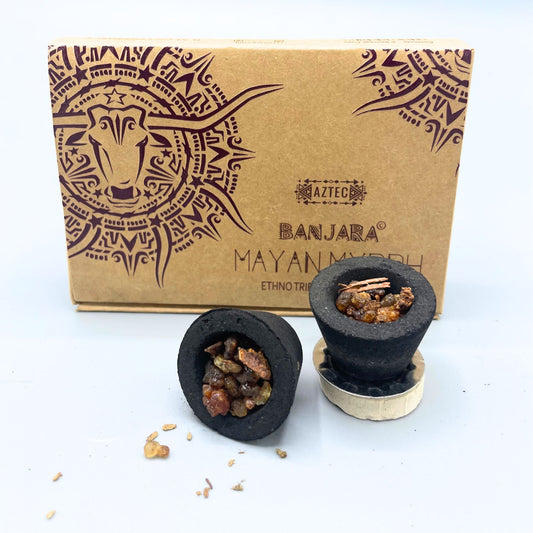 Incense cups hold resin chunks, placed on a metallic disc, in front of a cardboard box decorated with an intricate horned animal design. Text reads "AZTEC," "BANJARA," "MAYAN MYRRH," "ETHNO TRIBAL."