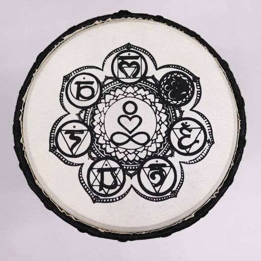 A circular drum features a central meditative symbol surrounded by intricate black mandala designs and chakra symbols on a white surface. The outer rim is textured, creating a harmonious aesthetic.