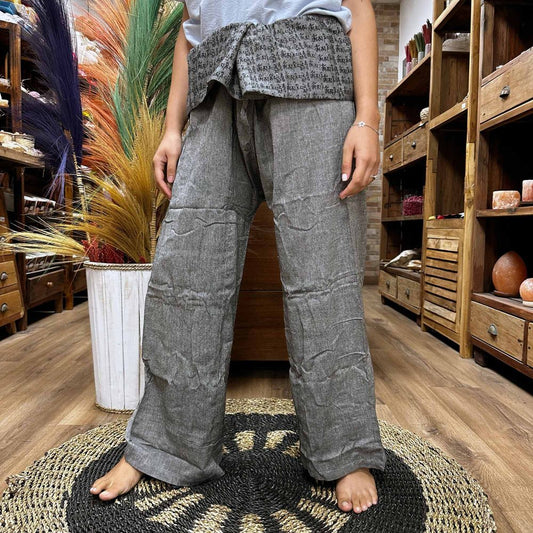 Gray Thai fisherman pants, standing on a woven rug, in a shop with wooden shelves.  The pants have a dark gray waistband with indecipherable text.