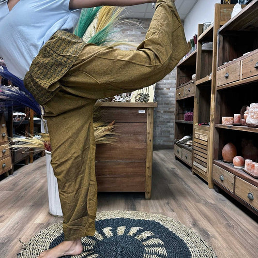 Mustard-colored pants with a patterned waistband are being modeled in a yoga pose.  The context is a shop with wooden shelving displaying various items.