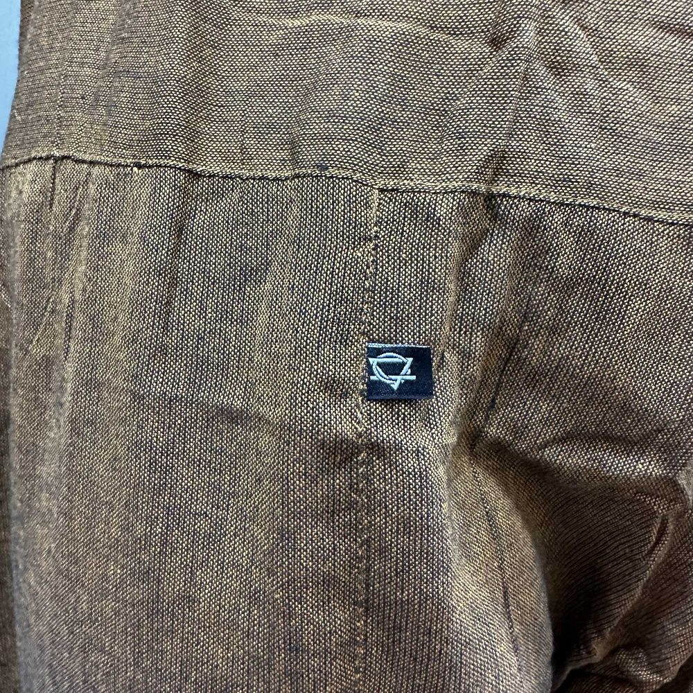 A brown, textured shirt back displays a small, rectangular black label with a stylized logo.