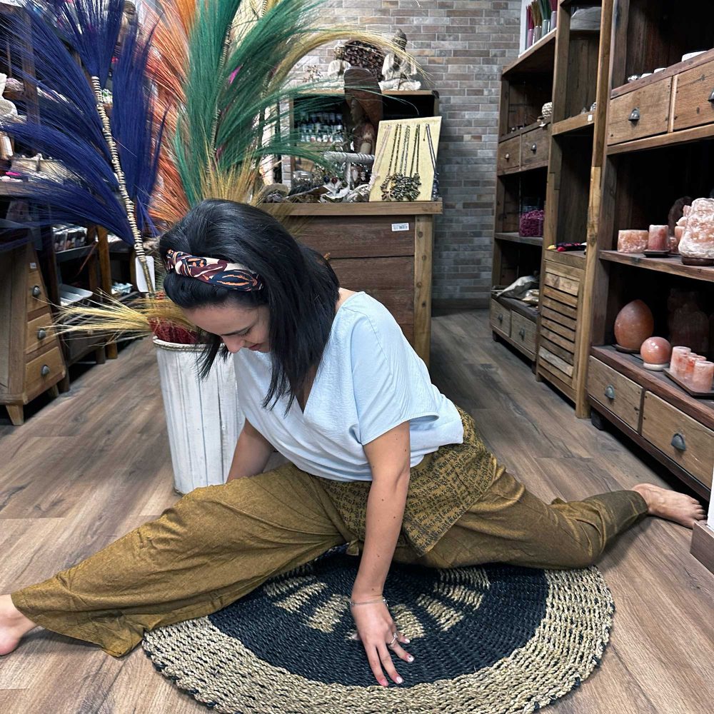 A woman, wearing a headband and loose pants, performs a seated leg stretch on a round mat. This is inside a shop with wooden shelves displaying various goods.