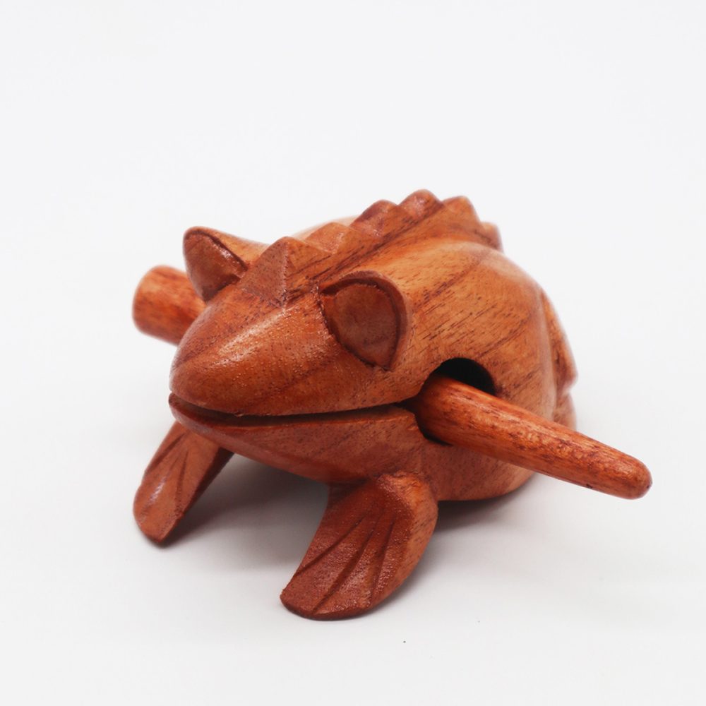 A wooden frog-shaped percussion instrument with a stick lodged in its mouth, placed on a smooth white surface. The frog features carved details, giving it a textured appearance.