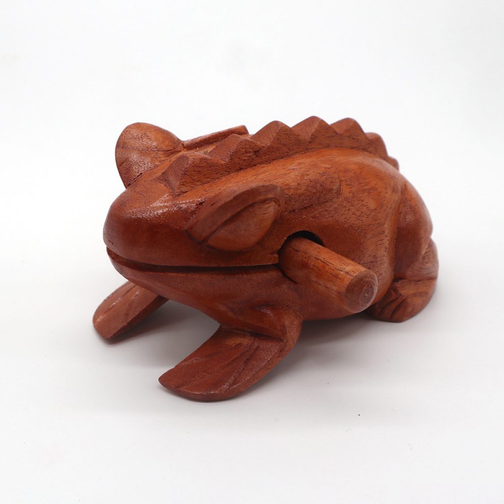 A wooden frog-shaped percussion instrument rests with a stick in its mouth, used to produce sound by rubbing along its ridged back. It is placed against a plain white background.