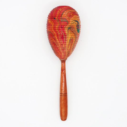 A colorful maraca with a textured, patterned head in red and orange hues, attached to a smooth wooden handle, is positioned against a plain white background.