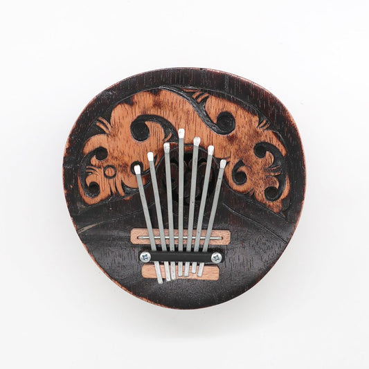 A wooden kalimba with seven metal tines is mounted on a carved, decorative surface with swirling patterns, placed on a plain white background.