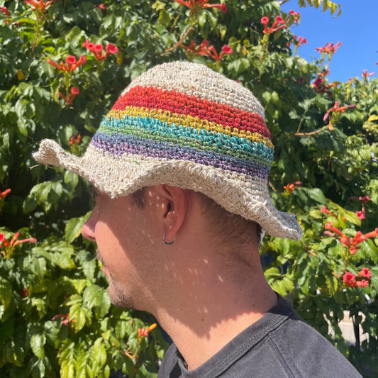 A crocheted hemp hat, featuring a rainbow band, is worn by a person, outdoors amidst flowering shrubs.