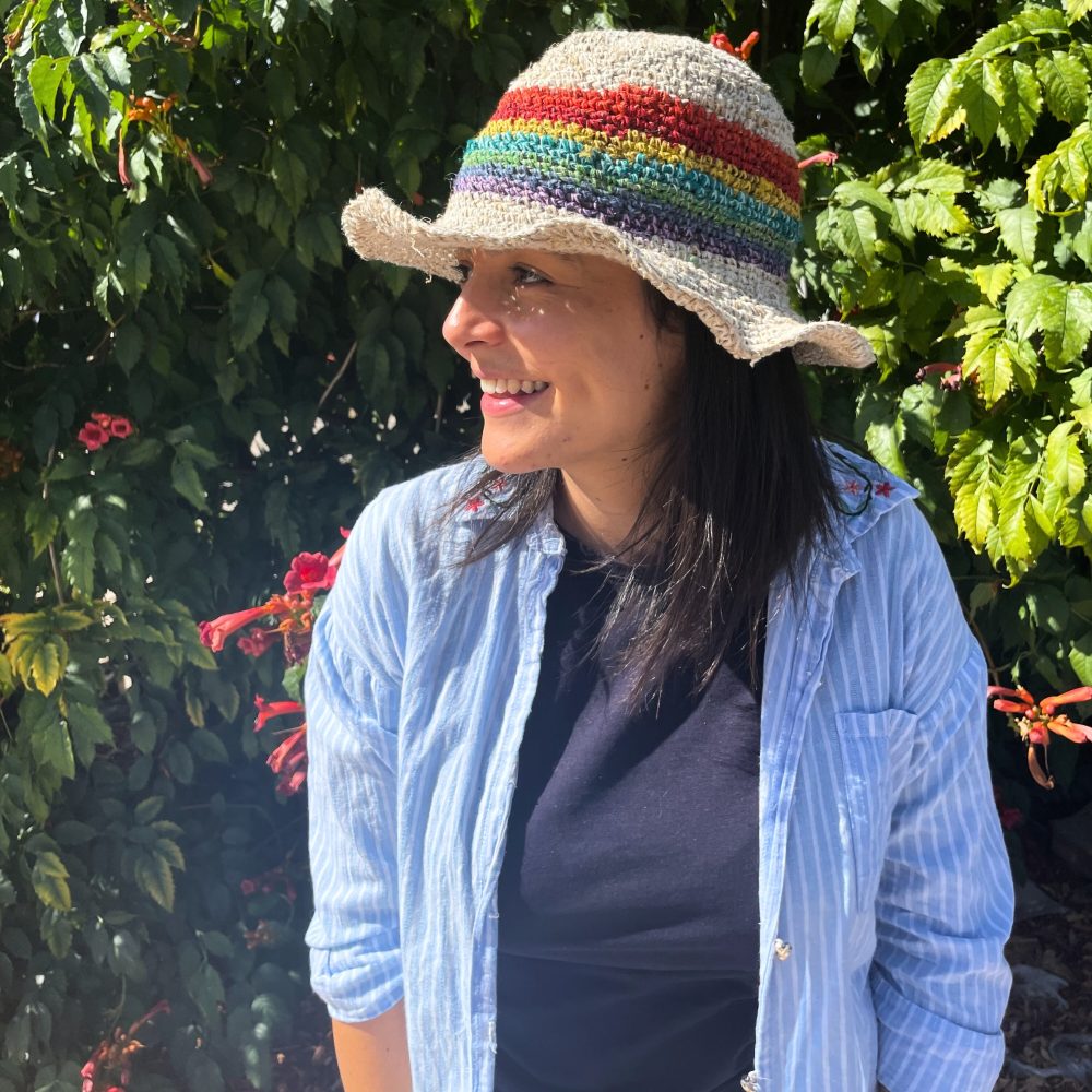 A crocheted sun hat with a rainbow band is worn by a woman. She smiles, standing outdoors amidst lush green foliage and reddish flowers.