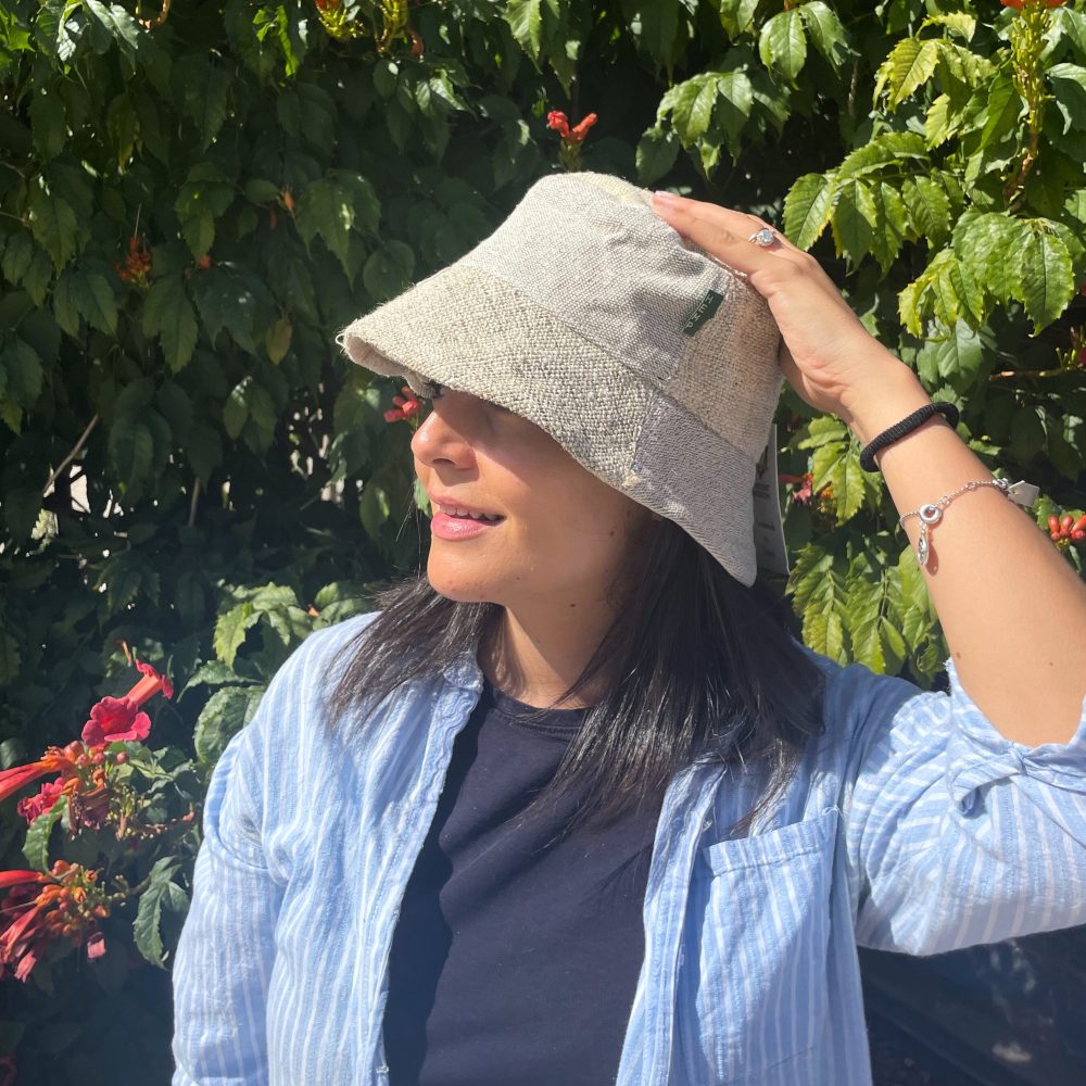 A woman adjusts a light beige bucket hat. She is outdoors, in front of green foliage with red flowers.