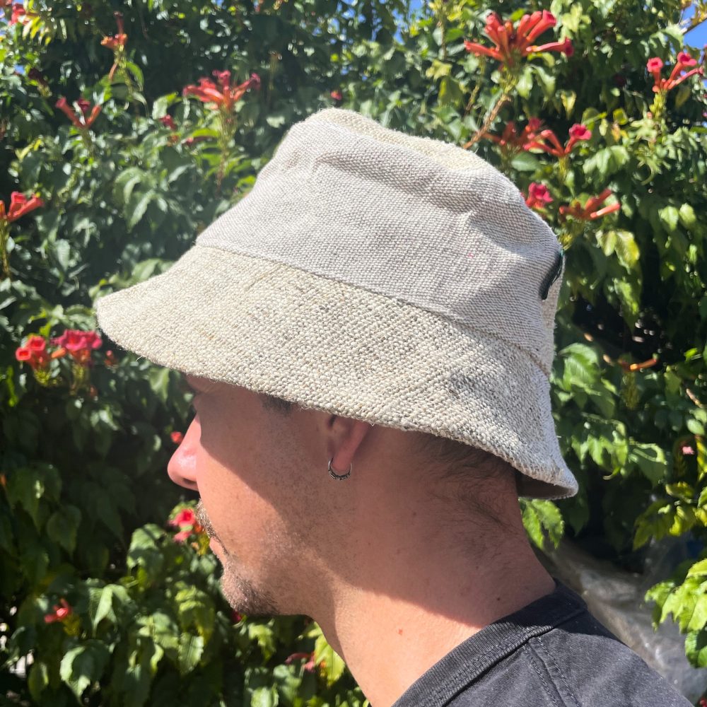 A hemp bucket hat is worn by a person, positioned in profile view against a flowering shrub backdrop.