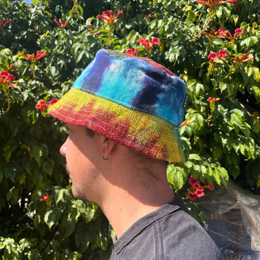 A tie-dye hemp bucket hat sits atop a person's head. The person is facing to the left, their profile visible against a backdrop of flowering green plants.