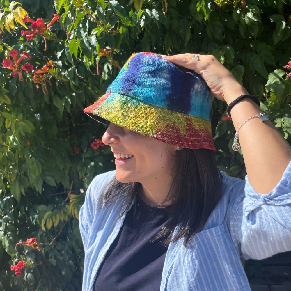 A tie-dye bucket hat is being adjusted by a woman.  She is smiling outdoors, near vibrant green foliage.
