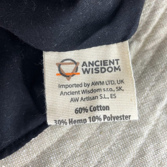 A fabric label displays “ANCIENT WISDOM,” material composition (60% cotton, 30% hemp, 10% polyester), and importer information (AWM LTD, UK; Ancient Wisdom s.r.o., SK; AW Artisan S.L., ES) attached to black fabric.