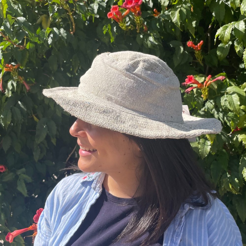 A light beige sun hat is worn by a person, who is smiling, in front of a flowering bush.