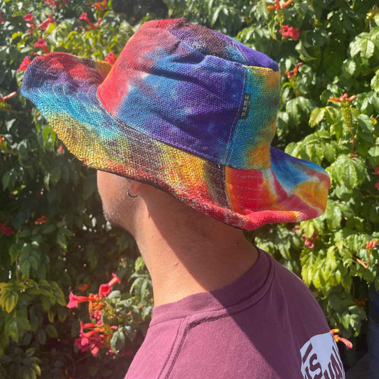A tie-dye hemp bucket hat sits atop a person's head outdoors. The hat displays a vibrant rainbow pattern. The person is wearing a maroon shirt and stands before a flowering bush. The hat's label reads "HEMP".