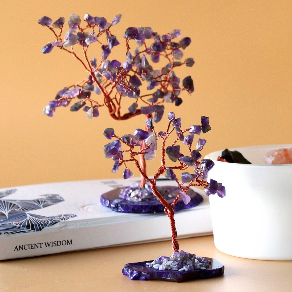Two amethyst wire-wrapped trees stand on agate bases atop a book titled "ANCIENT WISDOM". A white container is in the background.