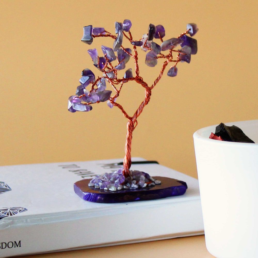 A copper wire tree, adorned with amethyst stones, sits on a white book. The base is also amethyst. A white container is visible in the background.