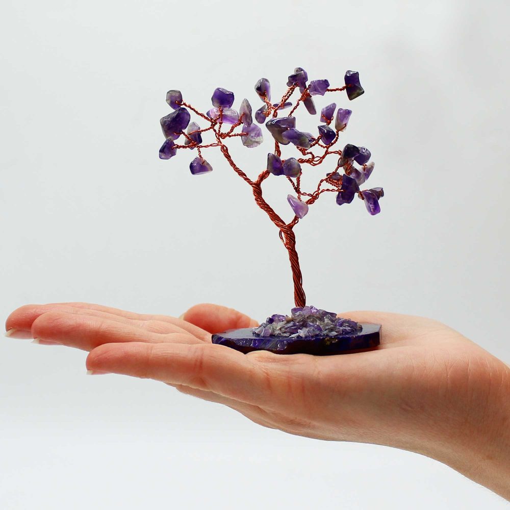 A small amethyst wire tree rests on an open palm. The tree is crafted from copper wire and purple amethyst stones. The background is plain white.