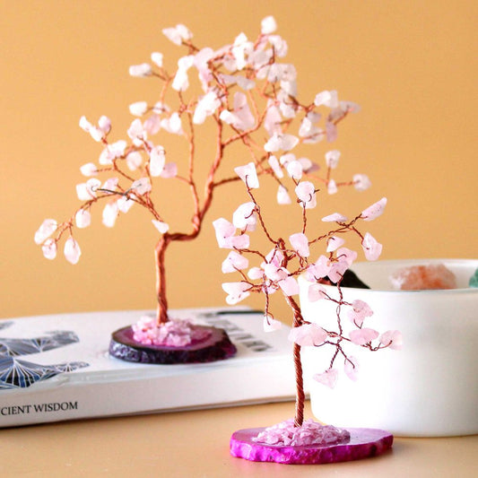 Two rose quartz wire-wrapped trees stand on agate bases atop a book; a container of crystals sits nearby.  "CIENT WISDOM" is partially visible on the book.