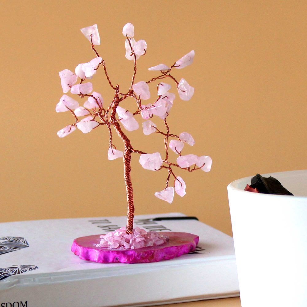 A rose quartz wire-wrapped tree stands on a fuchsia base atop a book. Pink crystals form the leaves; the trunk is copper wire. A white cup sits nearby.