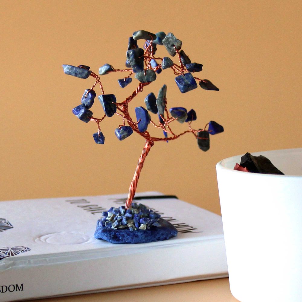 A lapis lazuli and copper wire tree stands on a book near a small white container. The tree's branches, crafted from wire, hold numerous small, blue stones.