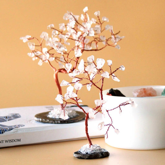 Two copper wire trees, adorned with clear quartz crystals, stand on a surface near a book and a white container.  "NT WISDOM" is visible on the book.