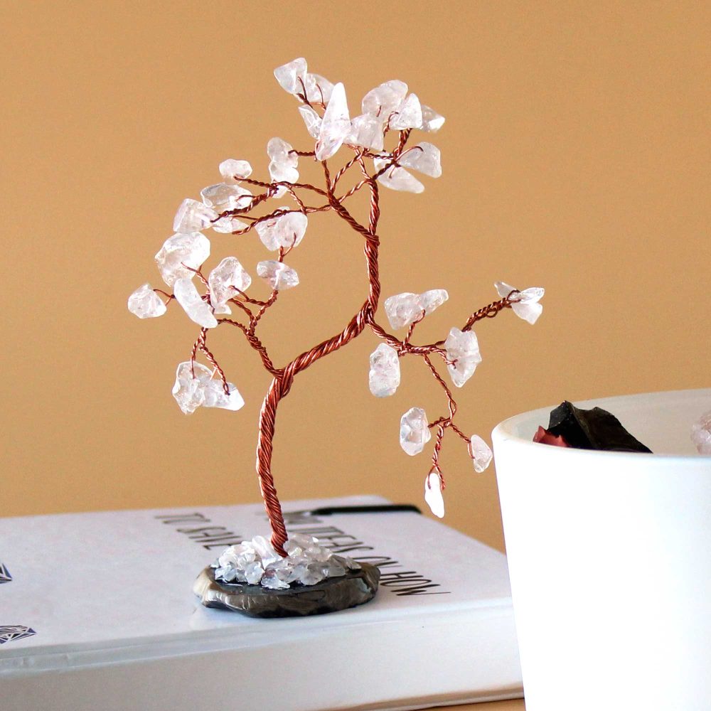 A copper wire tree, adorned with clear quartz crystals, sits on a book next to a white container.