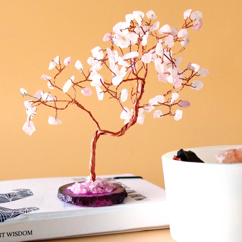 A rose quartz wire-wrapped tree stands on a book. The tree's base is a purple stone. It sits on a table near a white bowl. The visible text on the book is "NT WISDOM".