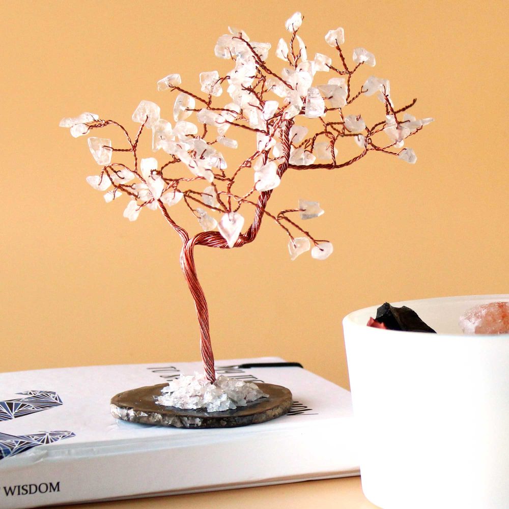 A copper wire tree, adorned with clear quartz crystals, sits on a book. The word "WISDOM" is visible on the book's cover. A white container is nearby.