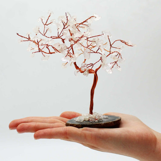A copper-wire tree, adorned with clear quartz crystals, rests on an agate slice; a hand gently supports it against a plain backdrop.